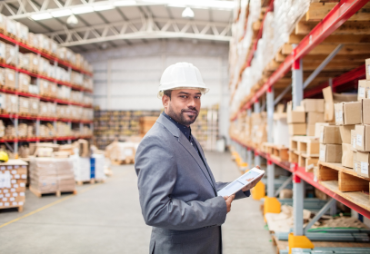 Man wearing a suit jacket and a hard hat holding a clipboard in a storage warehouse. He appears to be performing an audit.