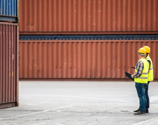 two men standing together looking at shipping containers while holding clipboards and wearing hard hats at a shipping and receiving dock.